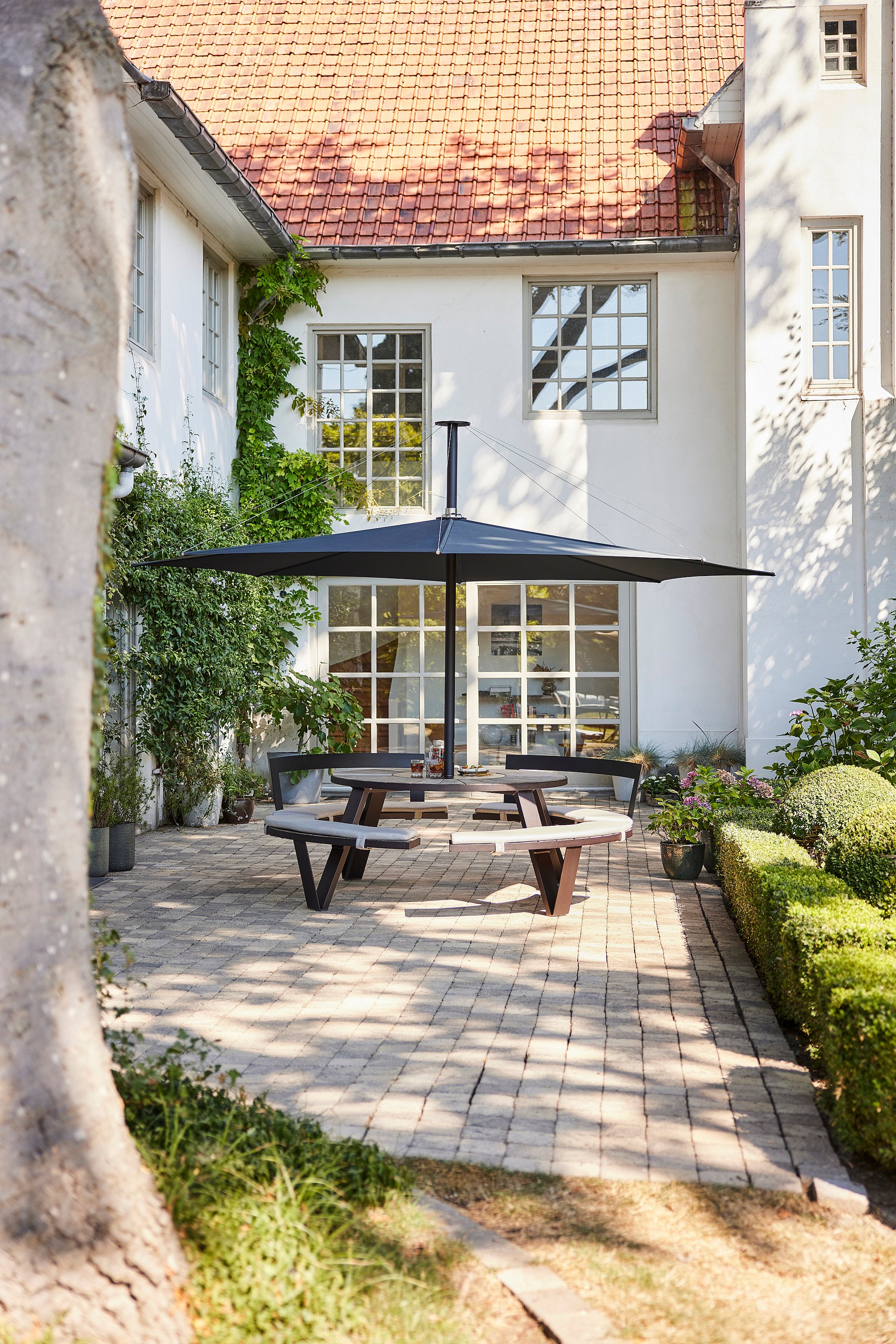 Vue d'ensemble d'une terrasse extérieure avec une table de pique-nique noire Extremis Pantagruel et un parasol Inumbra contrastant avec les murs blancs.