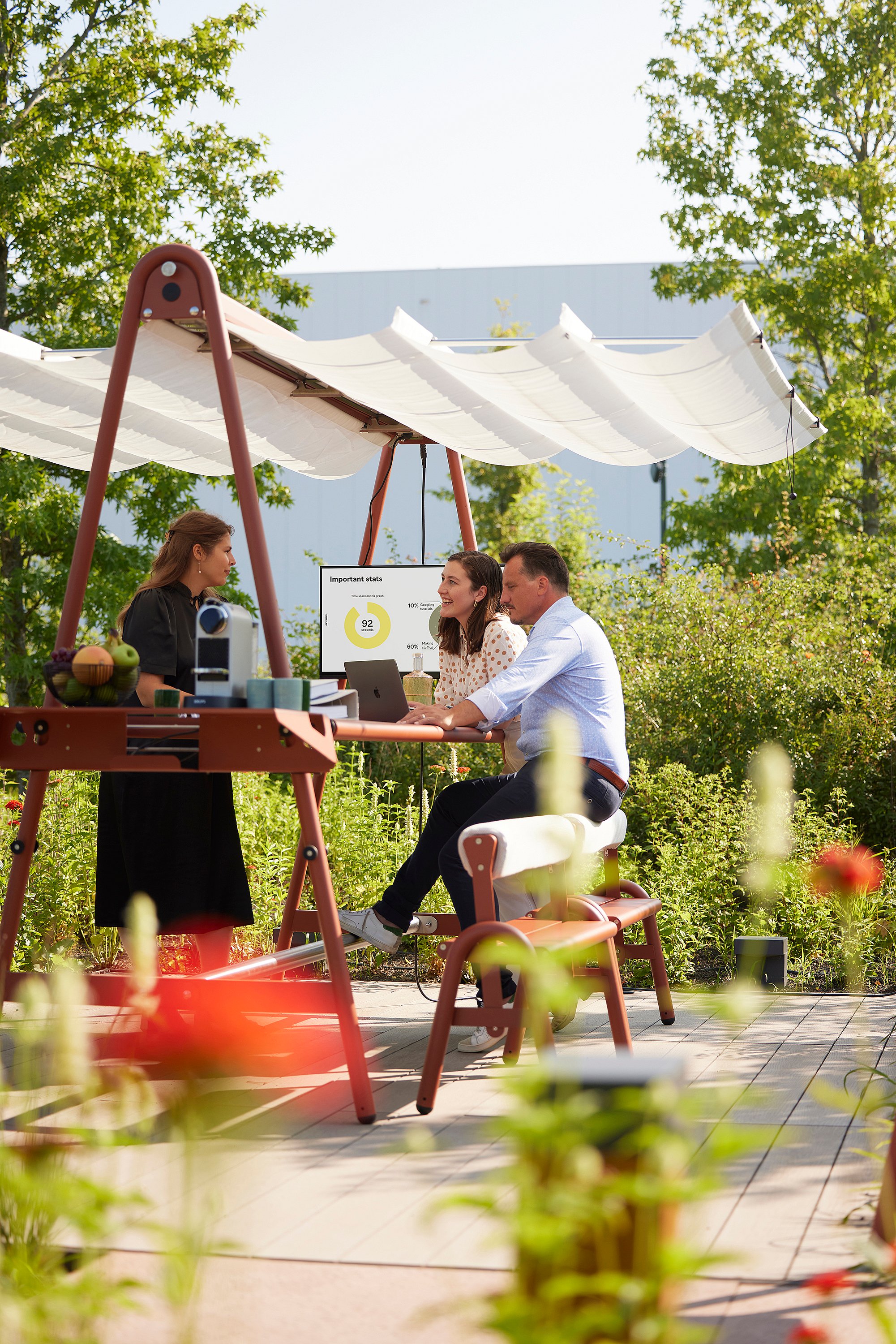 Drei Kollegen treffen sich an einem AMAi-Tisch im Freien unter einem Stoffdach; ein Laptop ist geöffnet und ein Präsentationsbildschirm steht in der Nähe in einer grünen Gartenumgebung.