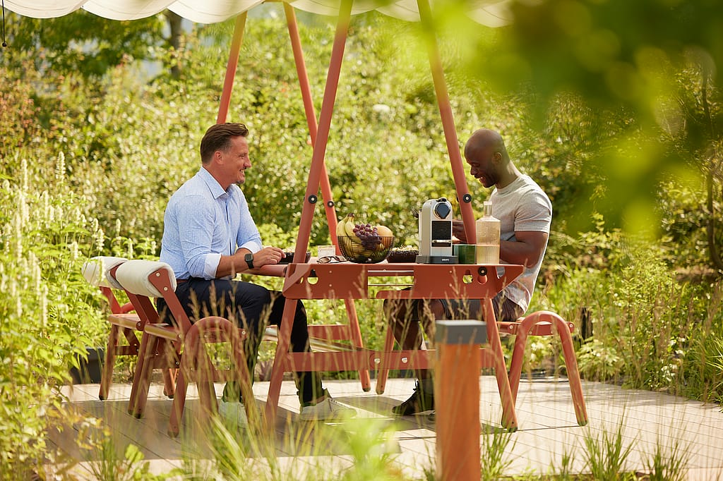 Zwei Kollegen sitzen sich an einem AMAi-Tisch im Freien gegenüber, auf dem eine Obstschale und eine Kaffeemaschine stehen, umgeben von Gartenpflanzen.