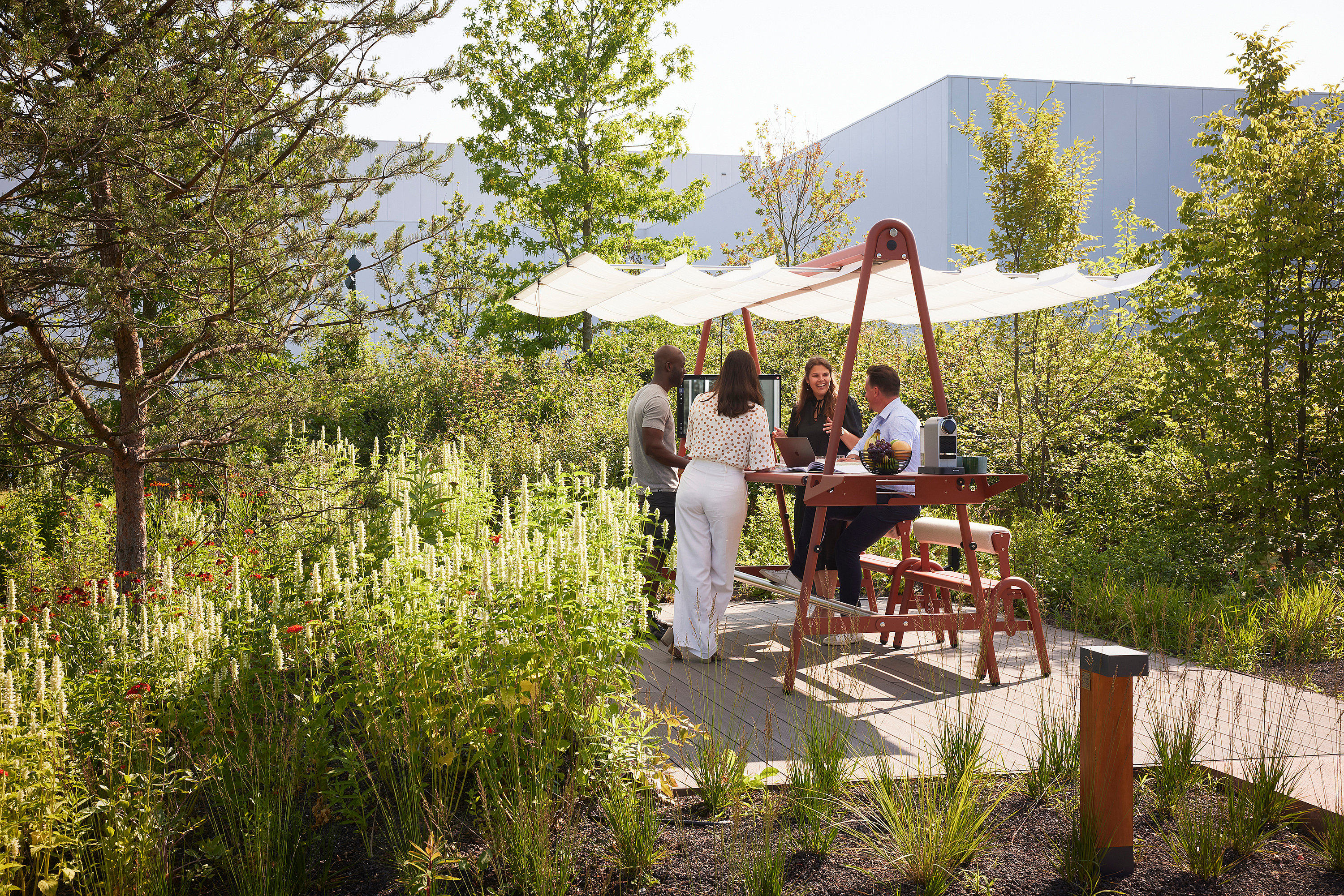 Vier Kollegen versammelten sich um einen AMAi-Tisch für Besprechungen im Freien mit Sonnenschutz und arbeiteten und unterhielten sich in einem begrünten Büro-Garten neben modernen Gebäuden.