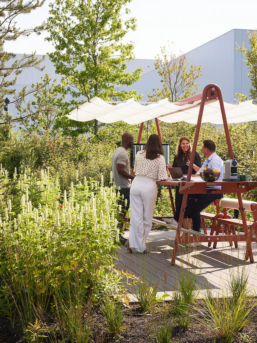 Four colleagues gathered around an AMAi outdoor meeting table with canopy shade, working and talking in a planted office garden beside modern buildings.