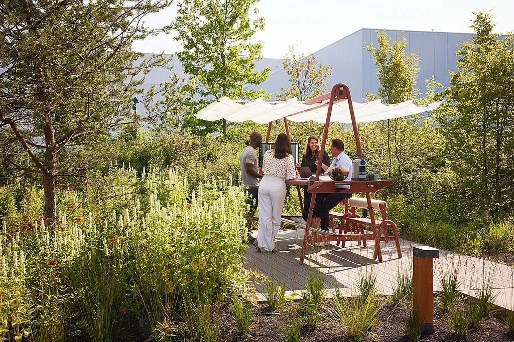 Vier Kollegen versammelten sich um einen AMAi-Tisch für Besprechungen im Freien mit Sonnenschutz und arbeiteten und unterhielten sich in einem begrünten Büro-Garten neben modernen Gebäuden.
