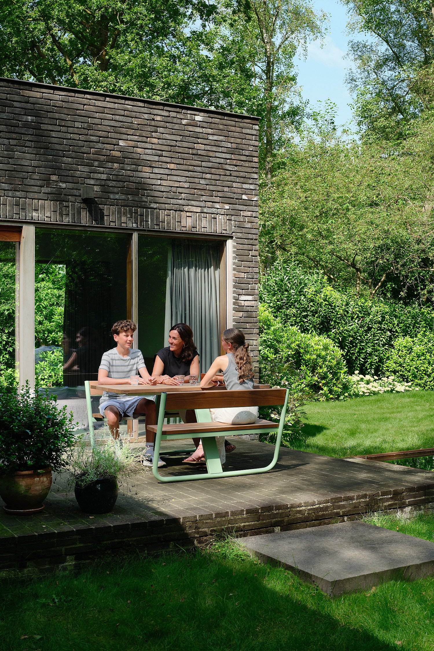 Ein grüner Picknicktisch „Manille“ mit integrierten Bänken steht auf einer kompakten Gartenterrasse neben einem Naturschwimmteich, umgeben von üppigen Bäumen und sanftem Sonnenlicht in Schilde, Belgien.