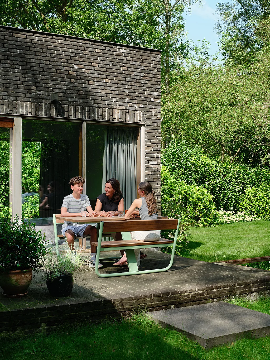 Ein grüner Picknicktisch „Manille“ mit integrierten Bänken steht auf einer kompakten Gartenterrasse neben einem Naturschwimmteich, umgeben von üppigen Bäumen und sanftem Sonnenlicht in Schilde, Belgien.