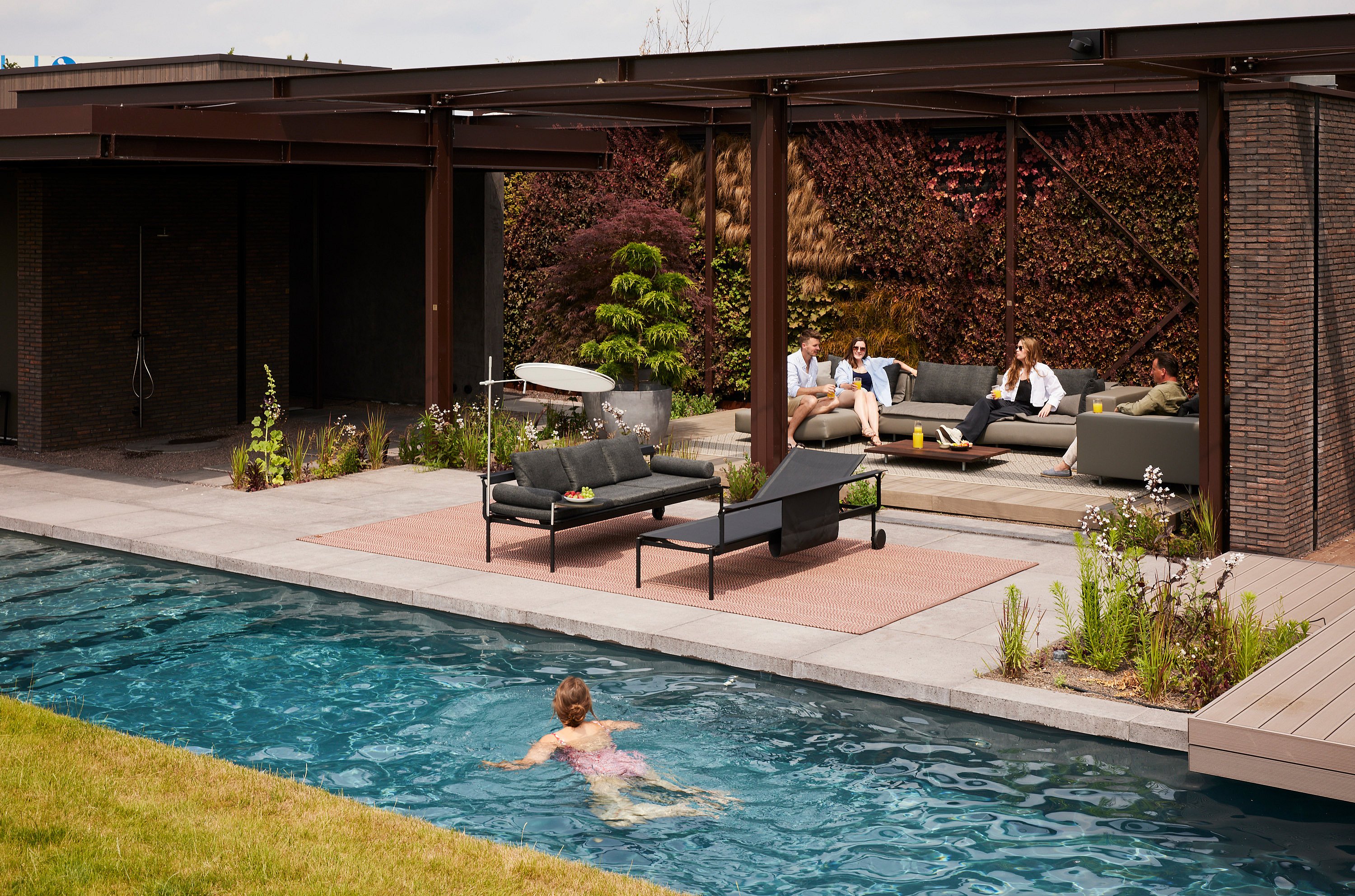   Eine sonnige Terrasse in Kruisem, Belgien, mit zwei Sol+Luna-Liegen neben einem Pool und einer Walrus-Lounge (Sofa, Stühle, Pouf und Beistelltisch) vor einer grünen Sichtschutzwand in warmen Rosttönen.