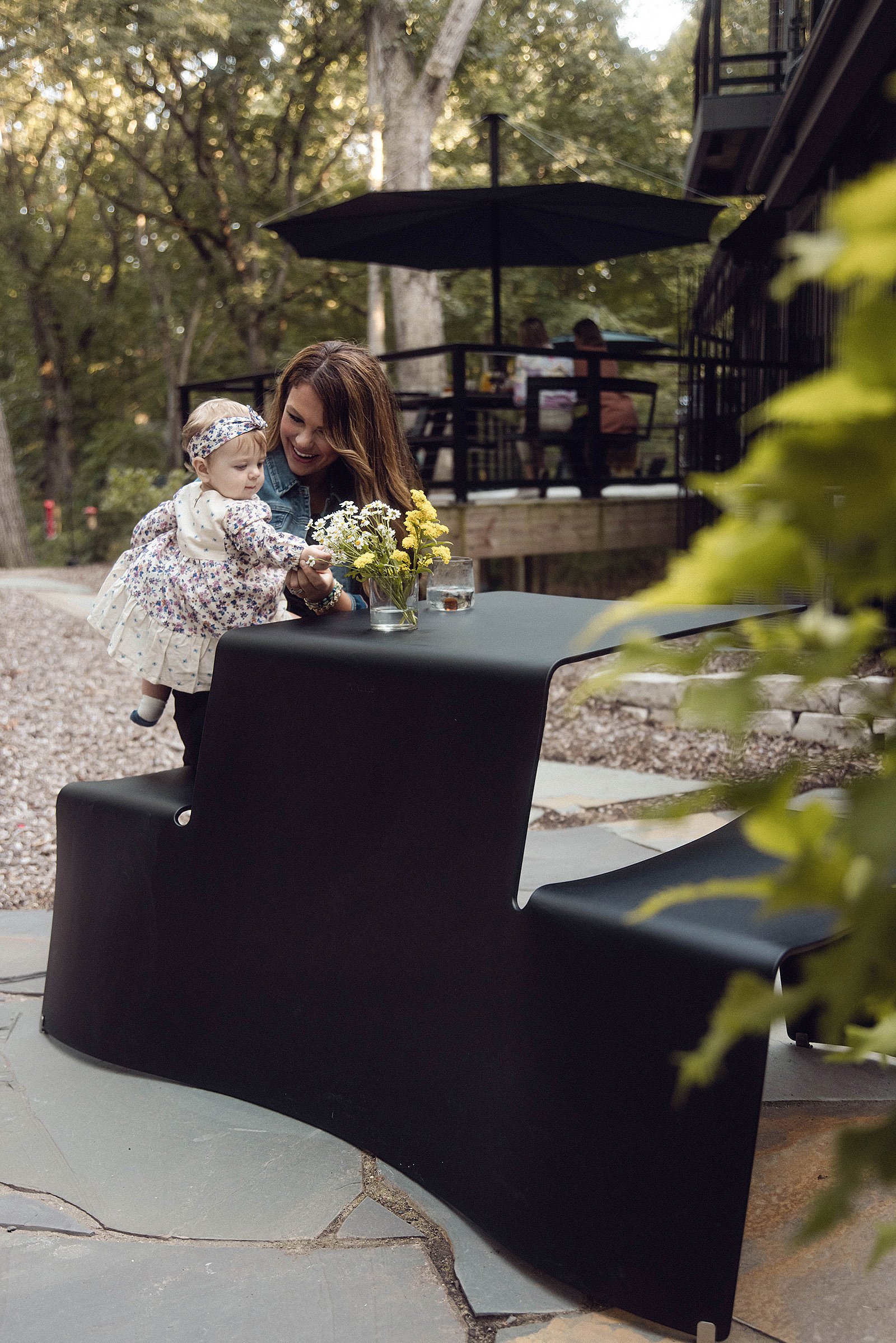 Une femme tenant un bébé sur une table de pique-nique avec bancs intégrés, sur une terrasse en pierre.