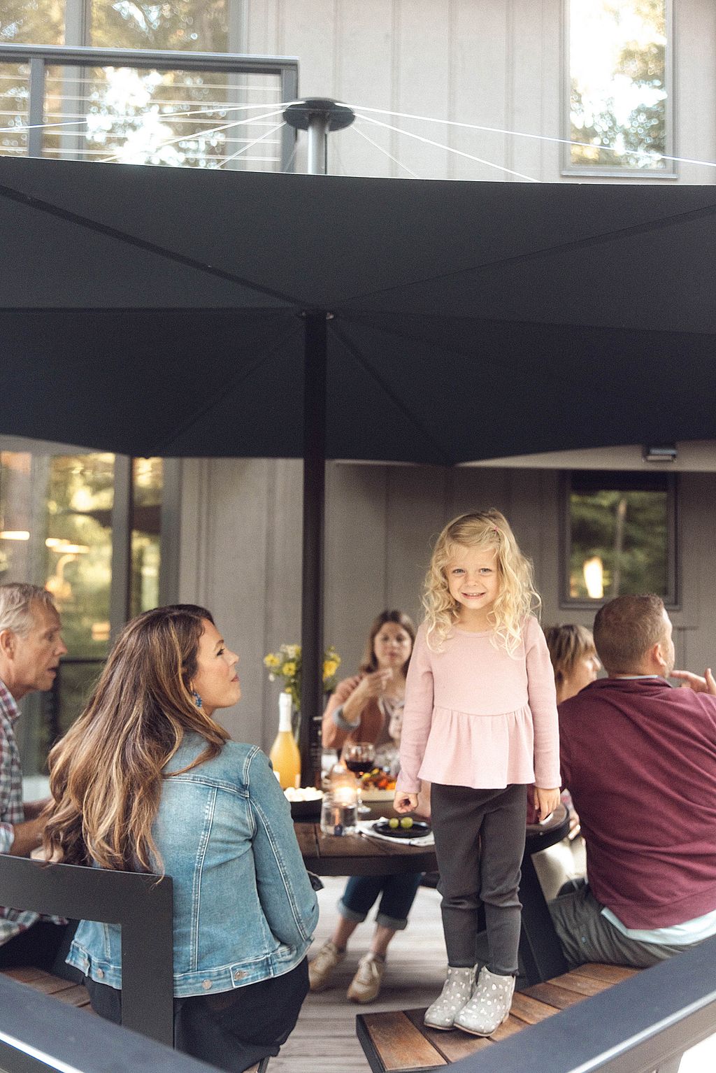 Famille réunie autour d'une table pantagruel sous un grand parasol noir Inumbra sur une terrasse en bois. Une jeune fille sourit sur un banc tandis que les adultes sont assis autour de la table avec des boissons et des amuse-gueules, créant une atmosphère chaleureuse et détendue.