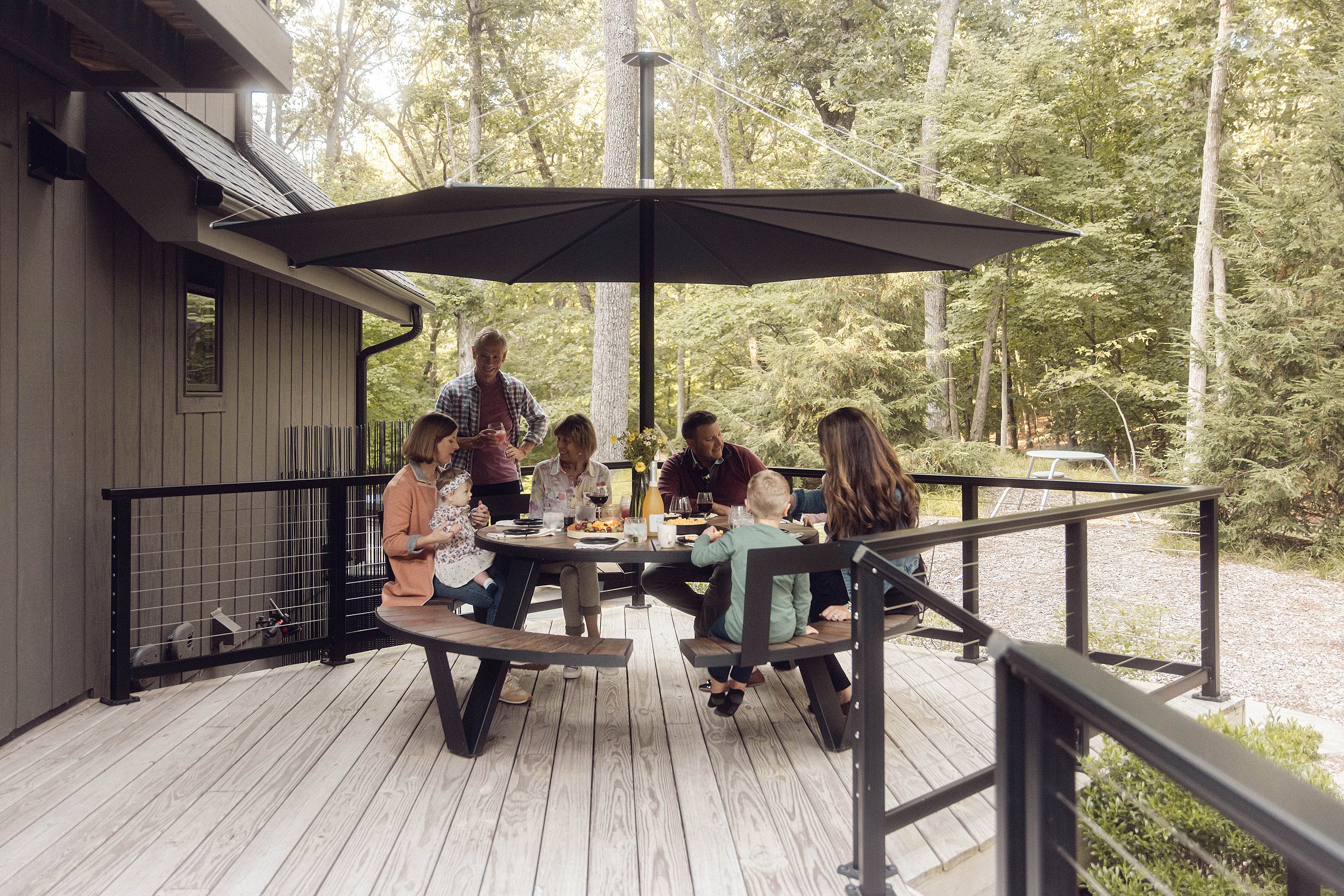 Vue d'ensemble d'une terrasse extérieure à Michiana Shores, avec plusieurs tables rondes et des bancs disposés sur une terrasse en bois clair. Des personnes sont assises et discutent dans les coins ombragés, entourées de pelouse verdoyante et de plantations.