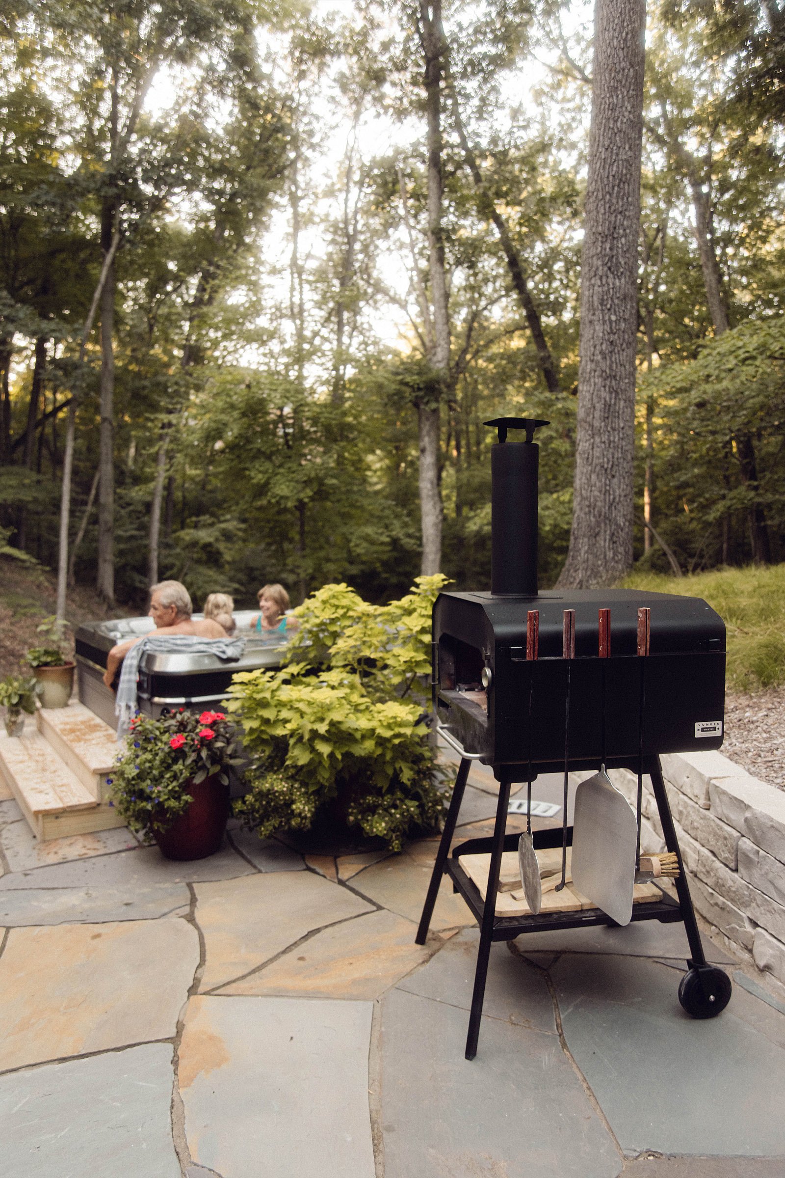 Four à pizza extérieur noir Vonken, avec cheminée et ustensiles suspendus, sur une terrasse en pierre dans un jardin boisé. À l'arrière-plan, trois personnes se détendent dans un jacuzzi entouré d'arbres, d'arbustes et de fleurs en pot