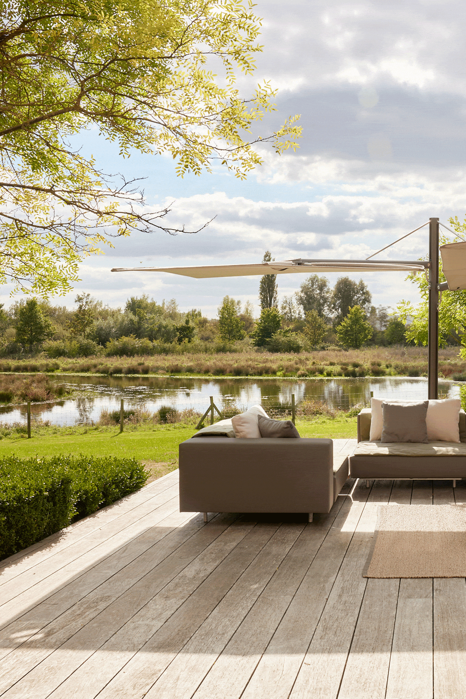 Beiger Sensu-Sonnenschirm, quadratisch, gerade, mit Rollengestell, spendet Schatten für ein Loungesofa auf einer Holzterrasse mit Blick auf einen Teich und Felder zur goldenen Stunde.