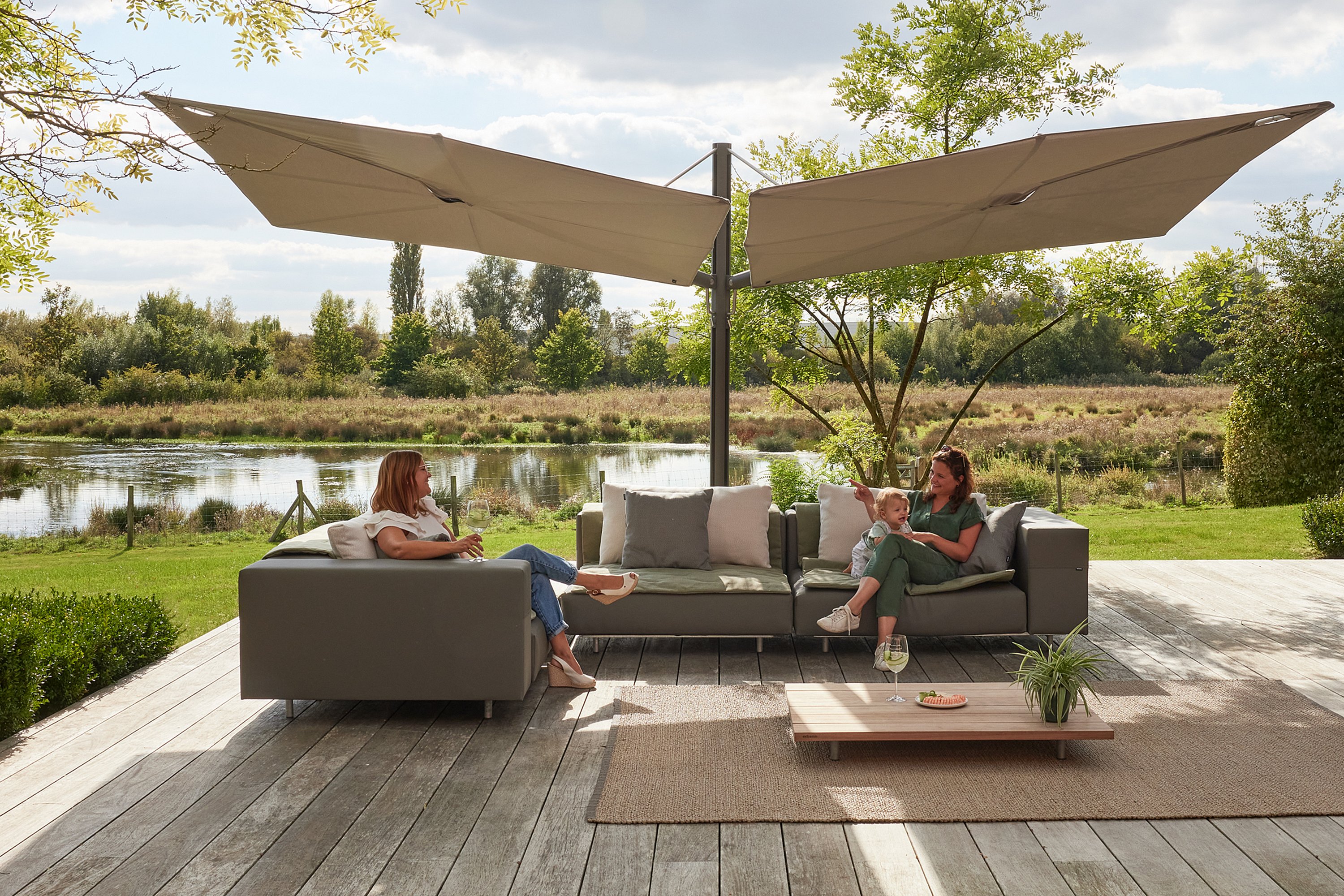 Terrasse extérieure avec un salon de jardin en forme de morse et deux grands parasols Sensu Duo Diamond fixés sur un mât central unique. Chaque toile est inclinée différemment pour créer une zone d'ombre superposée.