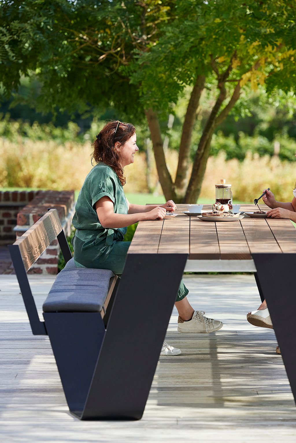 Nahaufnahme von Menschen, die an einem dunklen Hopper-Tisch auf einer Terrasse sitzen und Kaffeetassen, Kuchenteller und Desserts teilen.
