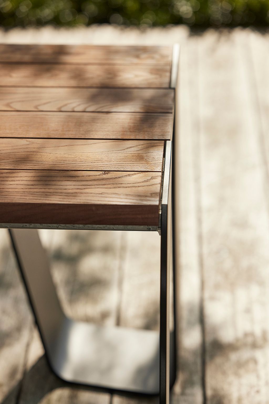 Nahaufnahme der hölzernen Tischplatte des Hopper-Tisches mit sichtbarer Maserung und Latten, getragen von einem schlanken Metallgestell. Fotografiert auf einer sonnigen Holzterrasse.