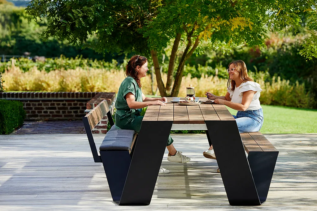 Outdoor terrace scene with a group of people seated around a long wooden Hopper table enjoying coffee and cake.
