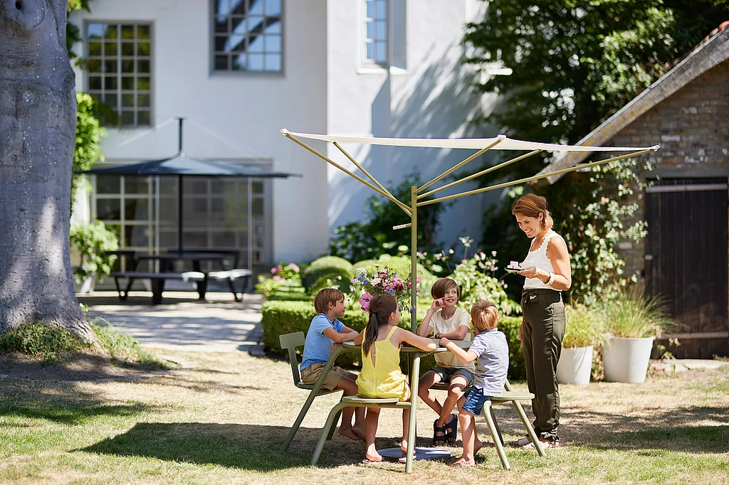 Outdoor garden scene with a large Virus picnic table set on a light stone patio. People sit and chat around the table while soft sunlight filters through surrounding trees and greenery.