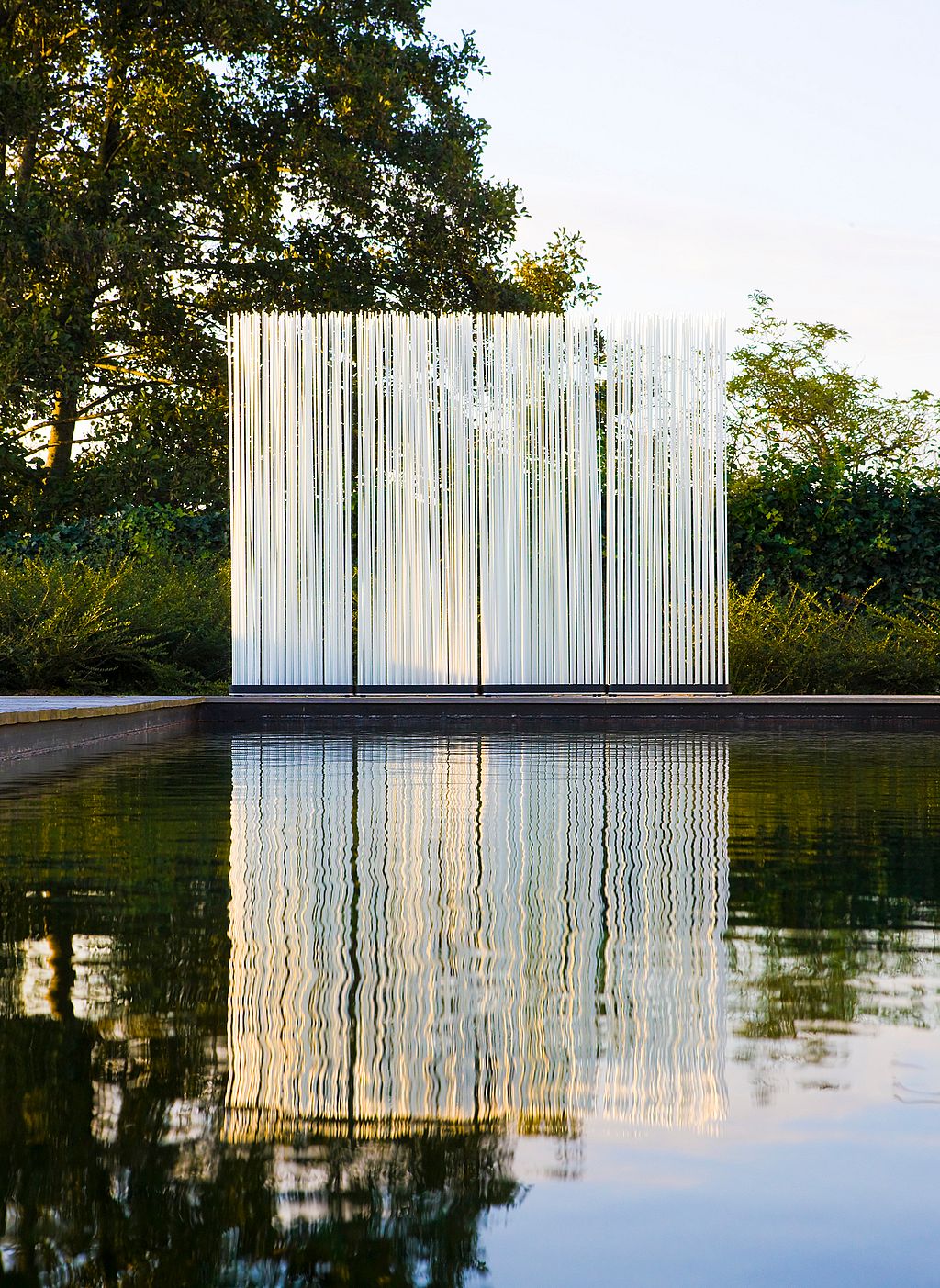 The Sticks outdoor lighting feature positioned beside a swimming pool at dusk. The vertical white illuminated rods form a semi-transparent screen, reflected in the still water, with trees and greenery in the background.