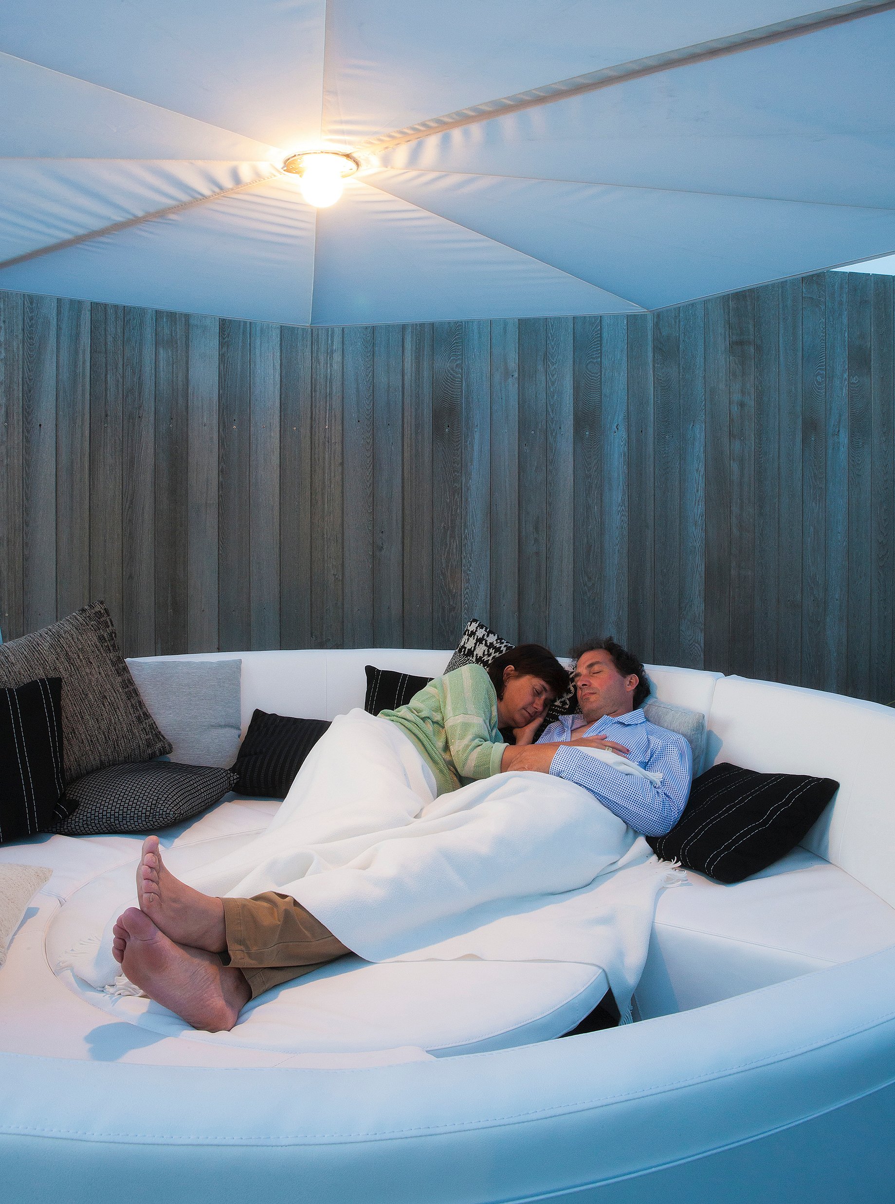 Ein Paar entspannt sich in der Abenddämmerung auf einem weißen Kosmos-Lounge-Sofa unter einem großen Sonnenschirm mit integrierter Beleuchtung.