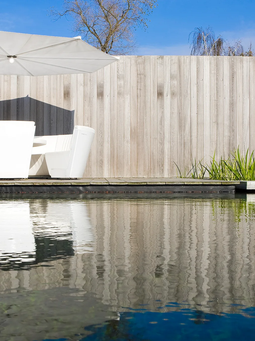 Een loungeset voor buiten, bestaande uit een comfortabele bank en een tafeltje, op een houten terras naast beplante borders en schaduwrijke plekjes.