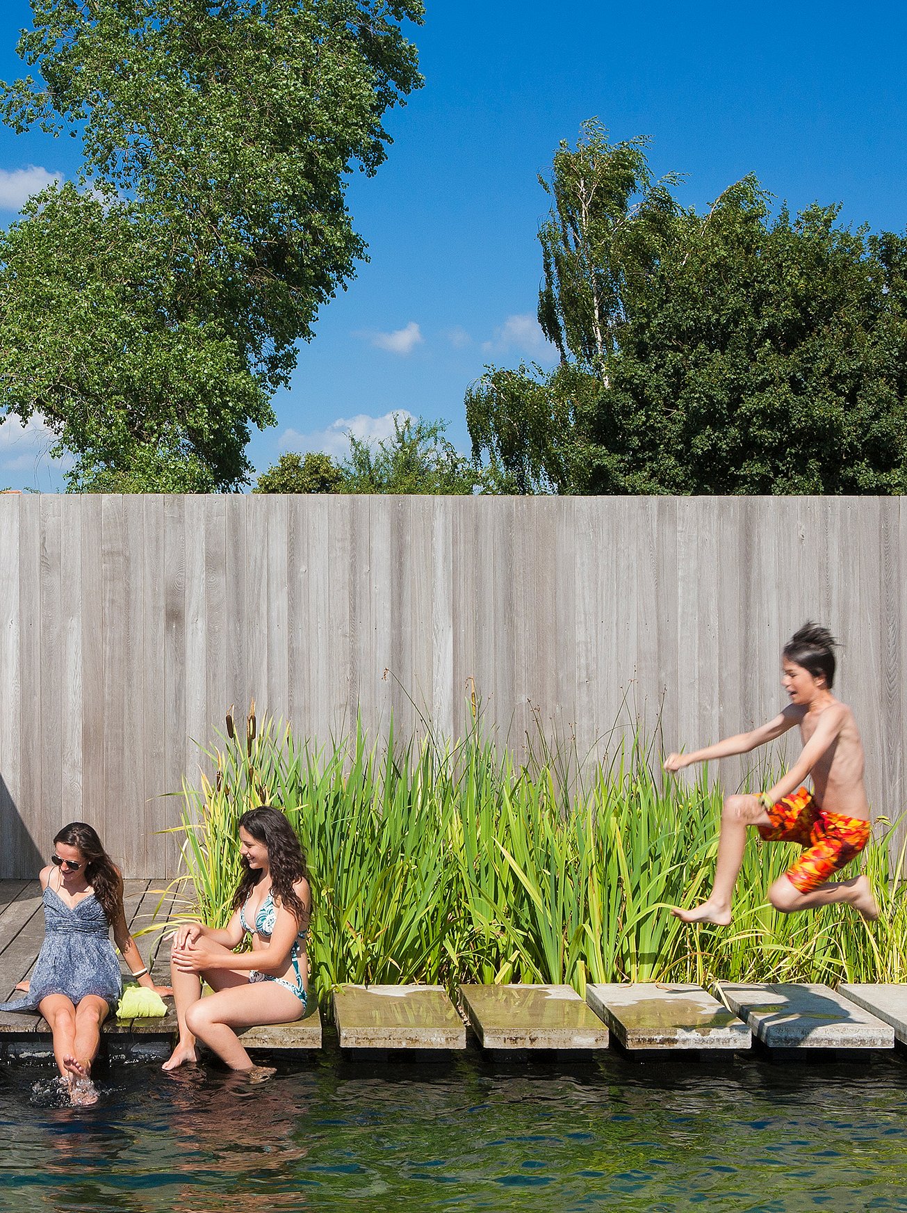 Zwei Mädchen sitzen am Rand eines Naturpools und baumeln mit den Füßen im Wasser, während ein Junge in bunten Badehosen von rechteckigen Trittsteinen ins Wasser springt.