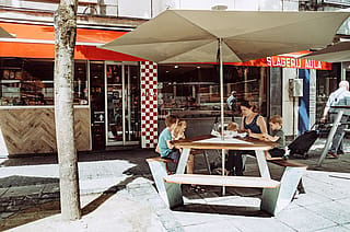 Adult and children seated at an outdoor picnic table under a parasol outside ‘Slagerij Aula’, with the shop entrance and red awnings behind them.