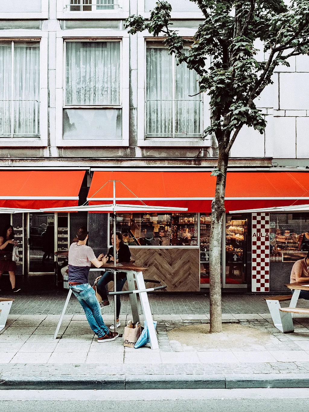 Terrasse de la boucherie Aula à Gand, avec deux tables de pique-nique Anker sous de grands parasols. Des clients déjeunent sous les auvents rouges vifs qui longent la façade.