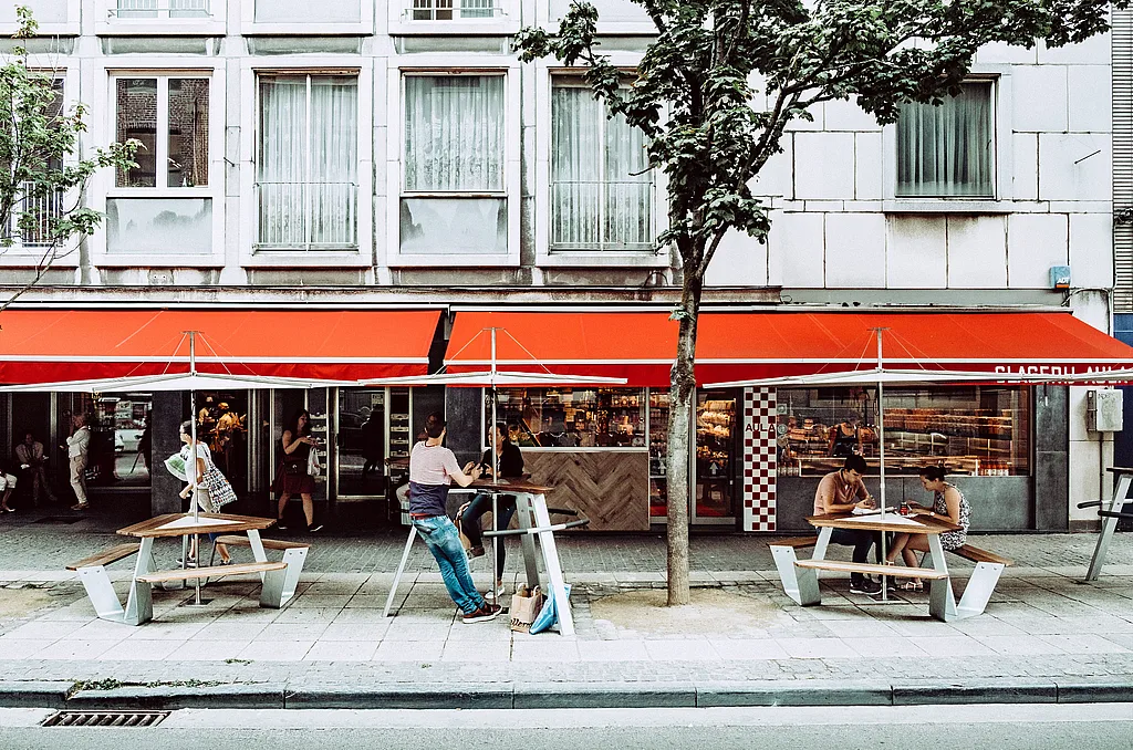 Street-side terrace outside butcher’s shop Aula in Ghent, with two Anker picnic tables under large parasols. People sit and eat beneath bright red awnings along the storefront.