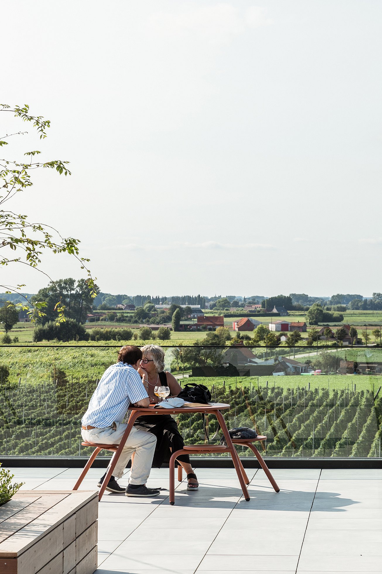 Tables extérieures dressées pour prendre un verre et grignoter au Bar Bernard sur son toit-terrasse. Des clients discutent sous des auvents tendus, avec vue sur les gratte-ciel urbains.