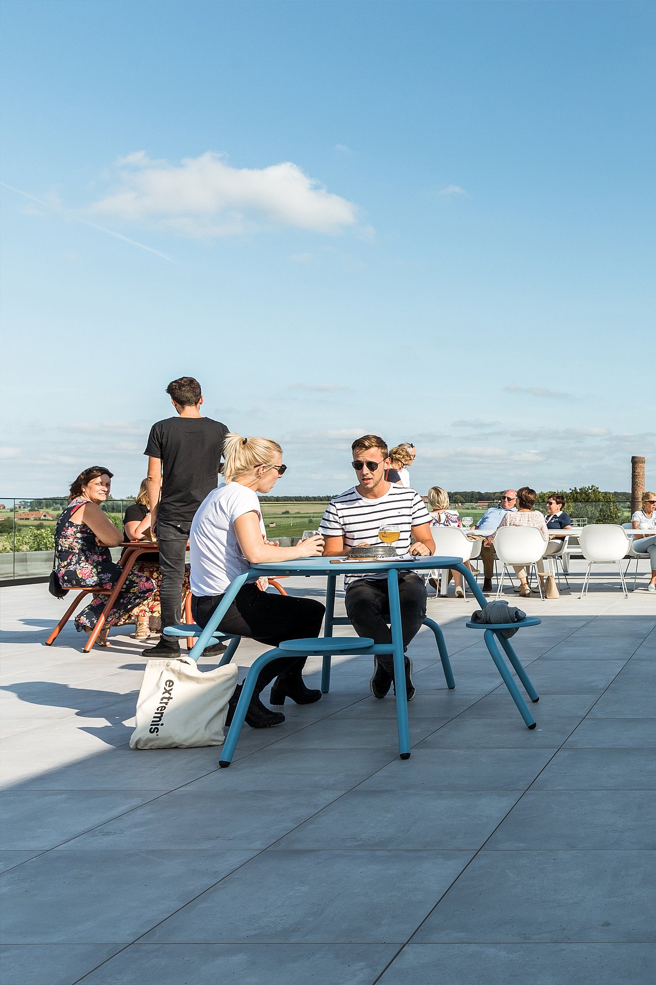 Des clients attablés sur la terrasse sur le toit-terrasse, à l'ombre du bâtiment. On aperçoit les immeubles et la silhouette de la ville en arrière-plan.