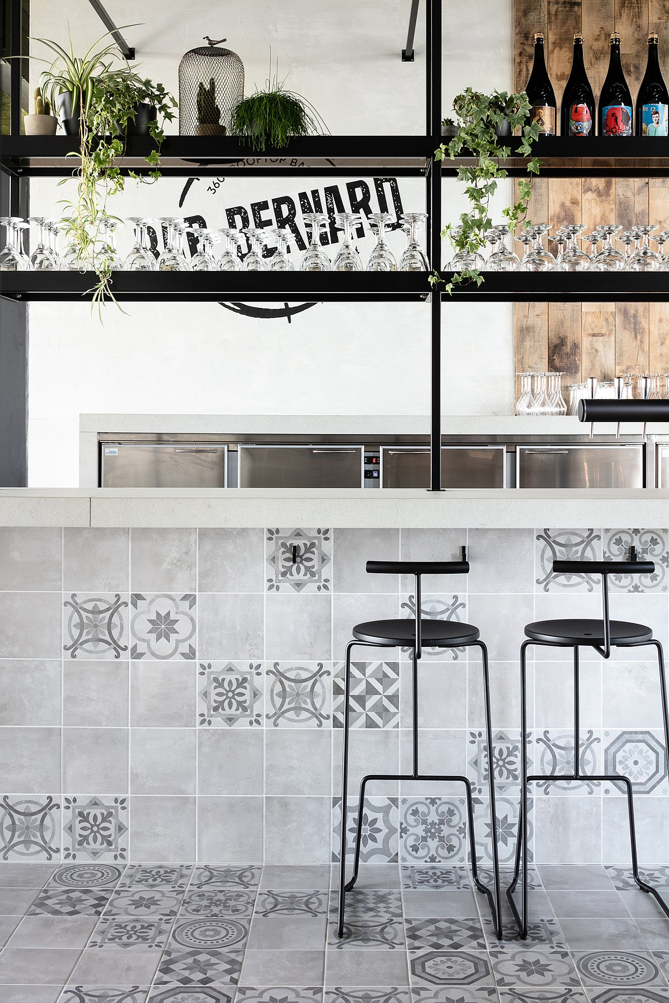 Intérieur moderne du bar : deux tabourets de bar en métal noir sont disposés autour d'un comptoir carrelé de carreaux de céramique gris et blancs à motifs. Au-dessus du comptoir, des étagères noires ouvertes présentent des verres à vin suspendus, des bouteilles et des plantes grimpantes, le tout sur un mur blanc orné du logo « Bar Bernard ».