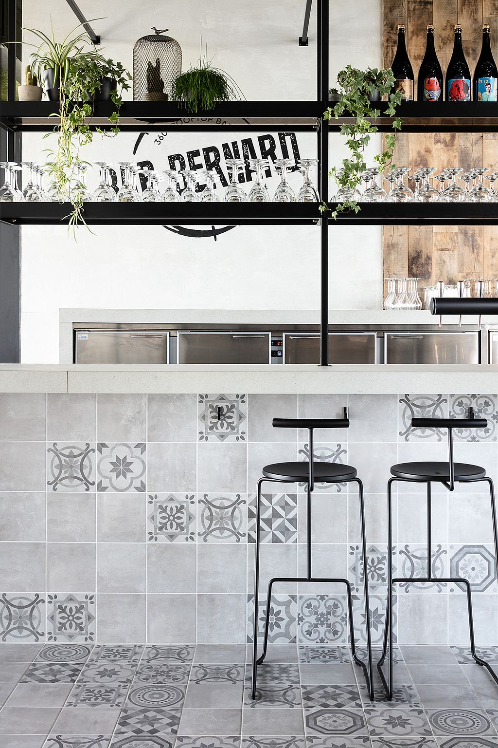Intérieur moderne du bar : deux tabourets de bar en métal noir sont disposés autour d'un comptoir carrelé de carreaux de céramique gris et blancs à motifs. Au-dessus du comptoir, des étagères noires ouvertes présentent des verres à vin suspendus, des bouteilles et des plantes grimpantes, le tout sur un mur blanc orné du logo « Bar Bernard ».