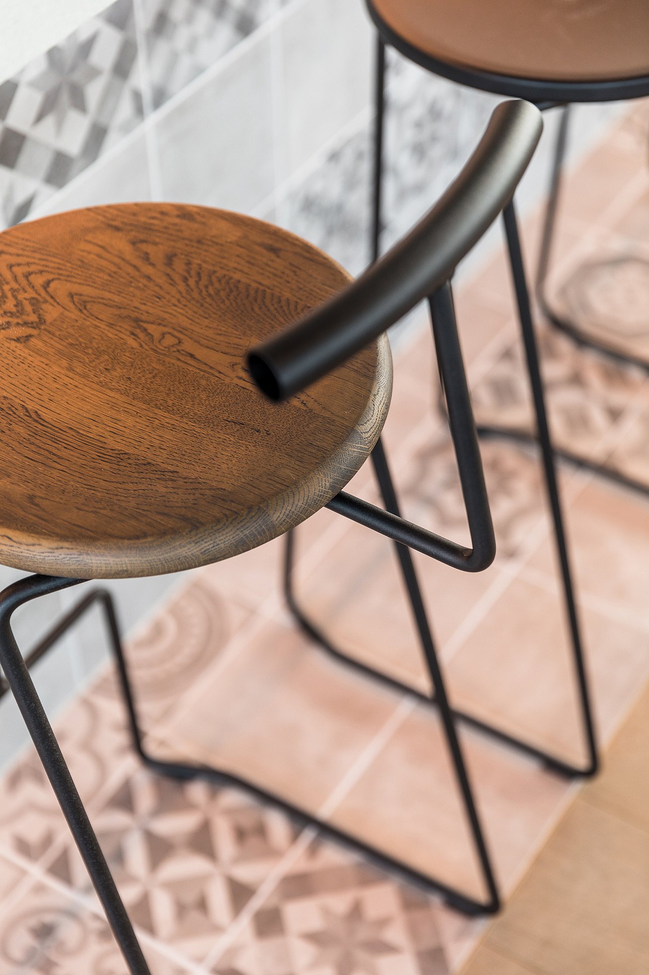 Gros plan sur un tabouret de bar : assise ronde en bois, structure en métal noir et dossier tubulaire incurvé. Le tabouret repose sur un sol carrelé de céramique à motifs.