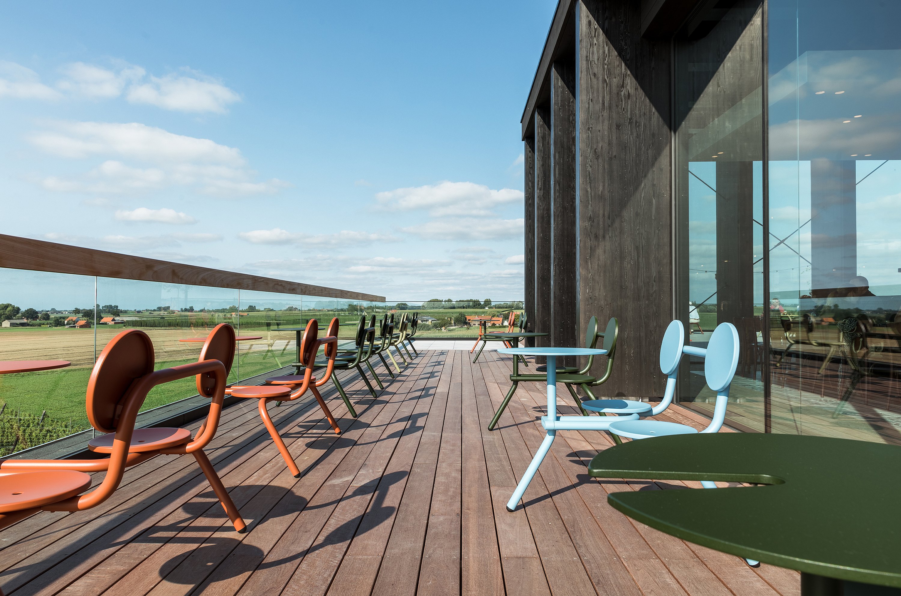 Dakterras met kleurrijke bistrotafels langs een glazen balustrade. Het houten terras biedt uitzicht op open velden onder een blauwe hemel.