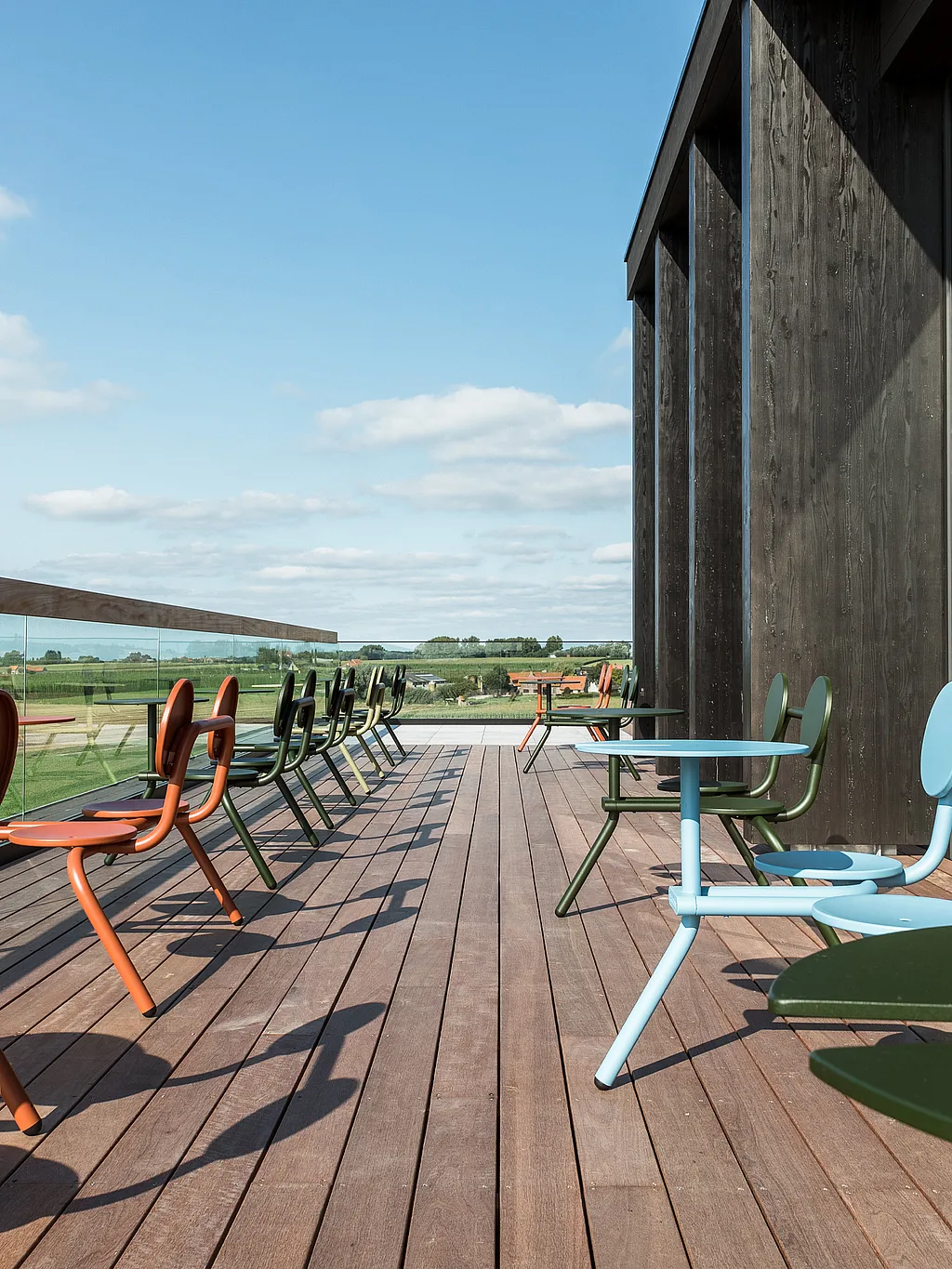 Dakterras met kleurrijke bistrotafels langs een glazen balustrade. Het houten terras biedt uitzicht op open velden onder een blauwe hemel.