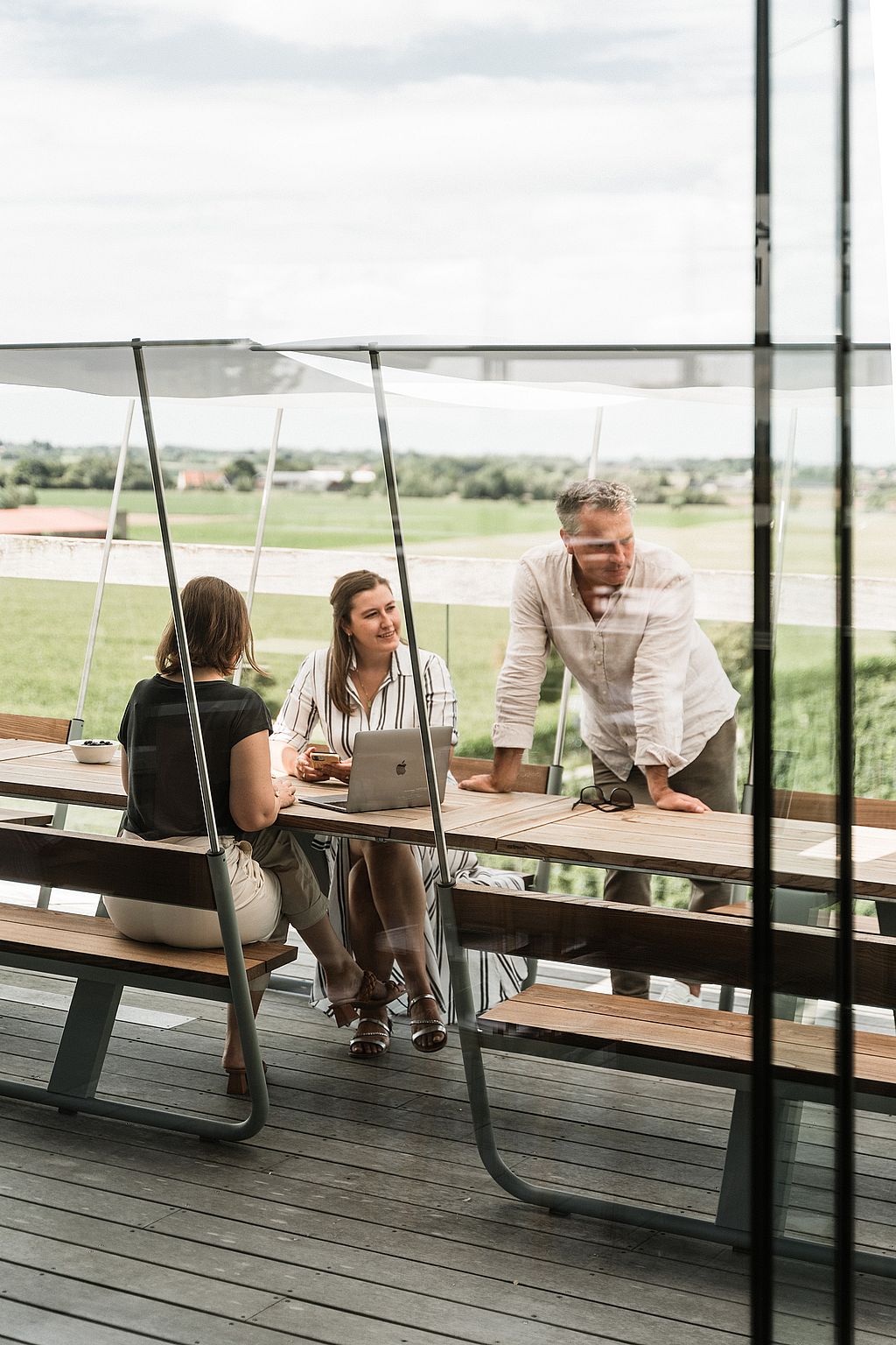 Several people work and talk at connected wooden Manille tables with integrated benches and white shade canopies on a terrace overlooking open countryside.