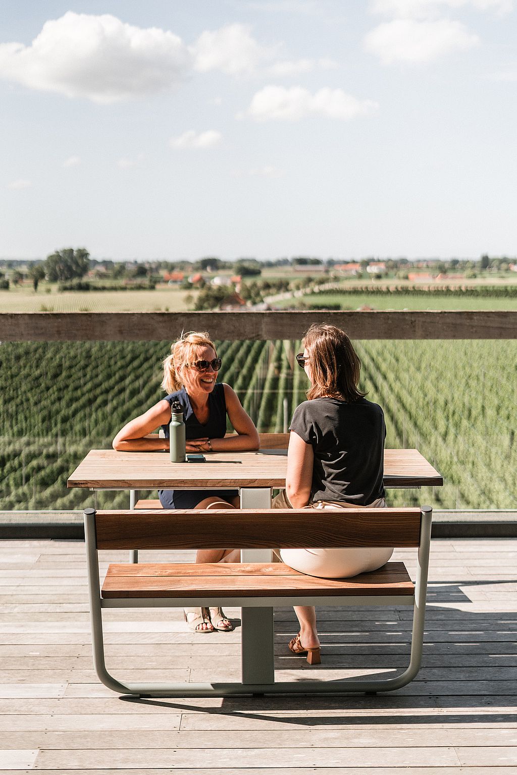 Two women sit and talk at a wooden Manille table on the terrace, with farmland stretching out behind the glass railing.