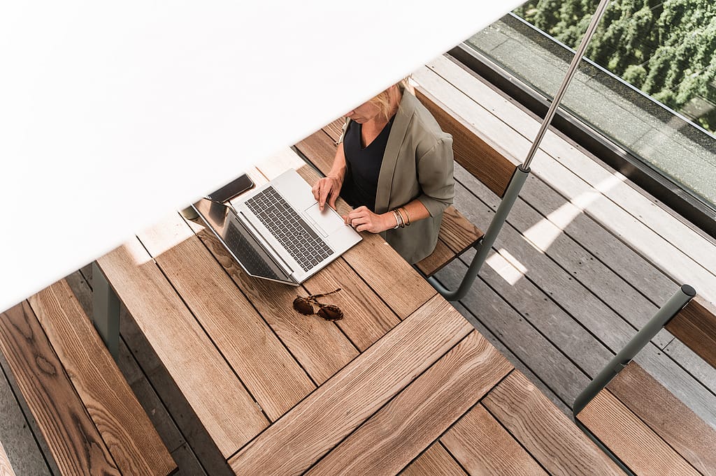 Top view of a woman working on a laptop at a wooden Manille table, partly shaded by the white canopy above.