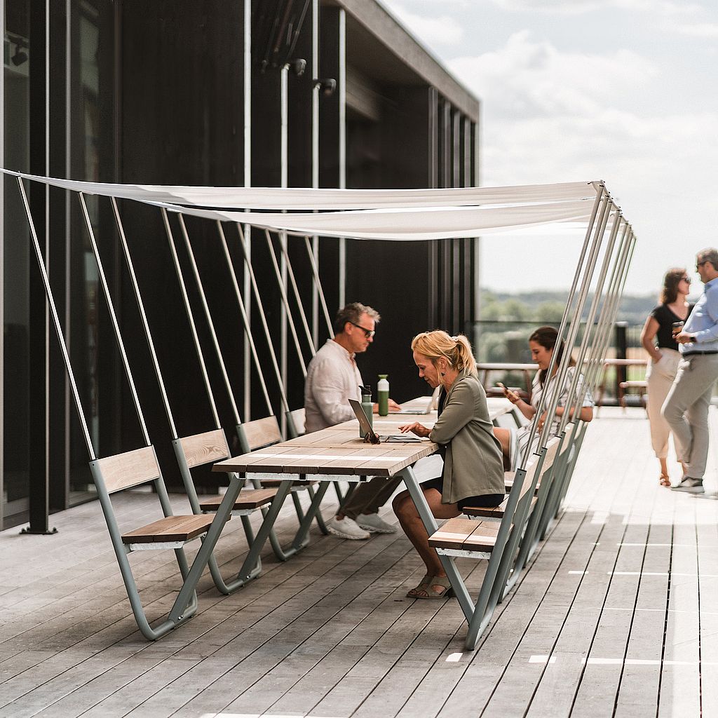People seated at a Manille picnic table with integrated shade canopy on a rooftop terrace, with a city view beyond.