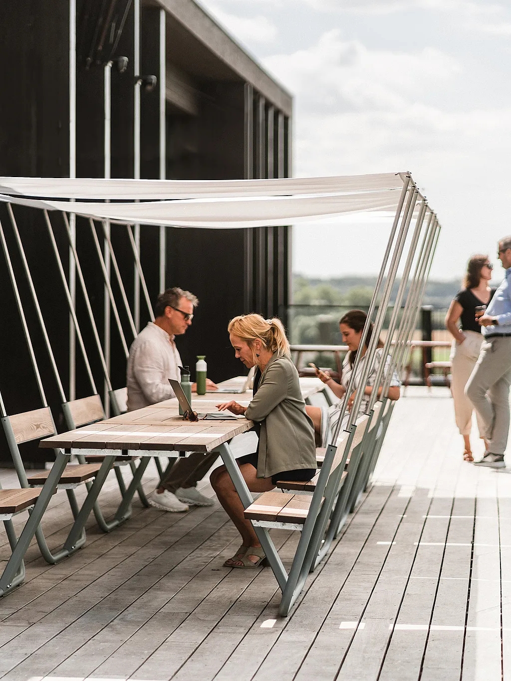Mehrere Personen arbeiten und unterhalten sich an verbundenen hölzernen Manille-Tischen mit integrierten Bänken und weißen Schattenüberdachungen auf einer Terrasse mit Blick auf die offene Landschaft.