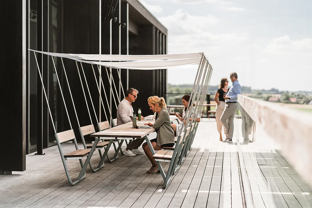 Several people work and talk at connected wooden Manille tables with integrated benches and white shade canopies on a terrace overlooking open countryside.