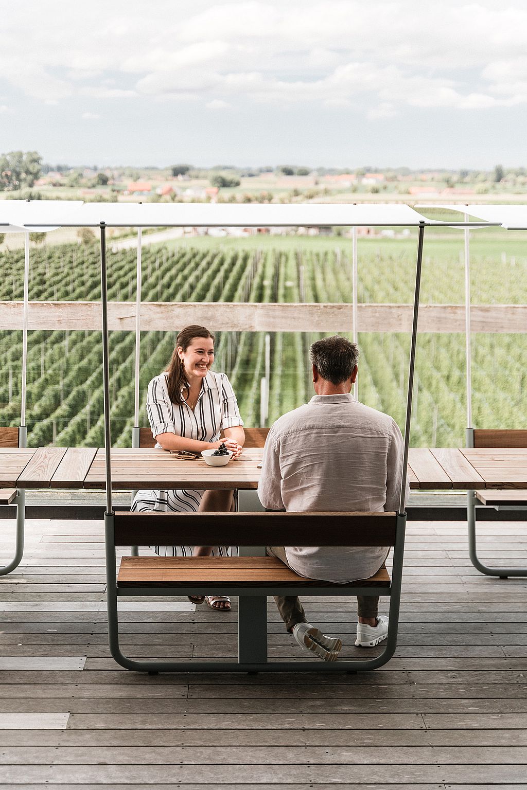 Two people sit opposite each other at a wooden Manille table with a white canopy, framed by a glass balustrade and neat rows of fields beyond.