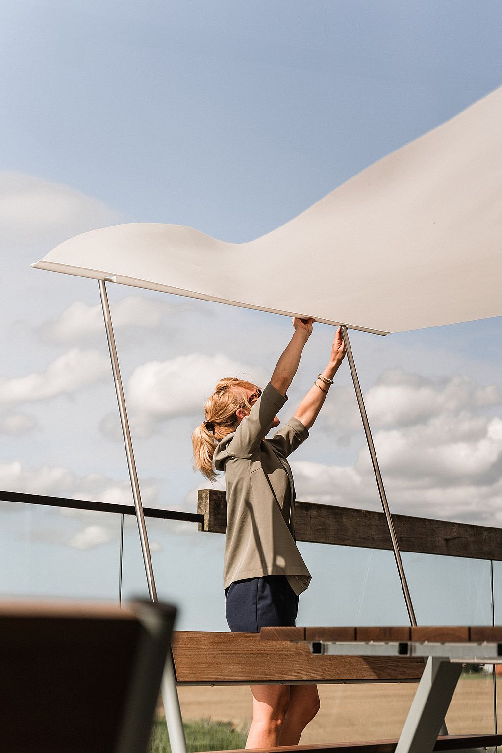 A woman raises the white fabric shade above a Manille table on the terrace, with open sky and fields in the background.