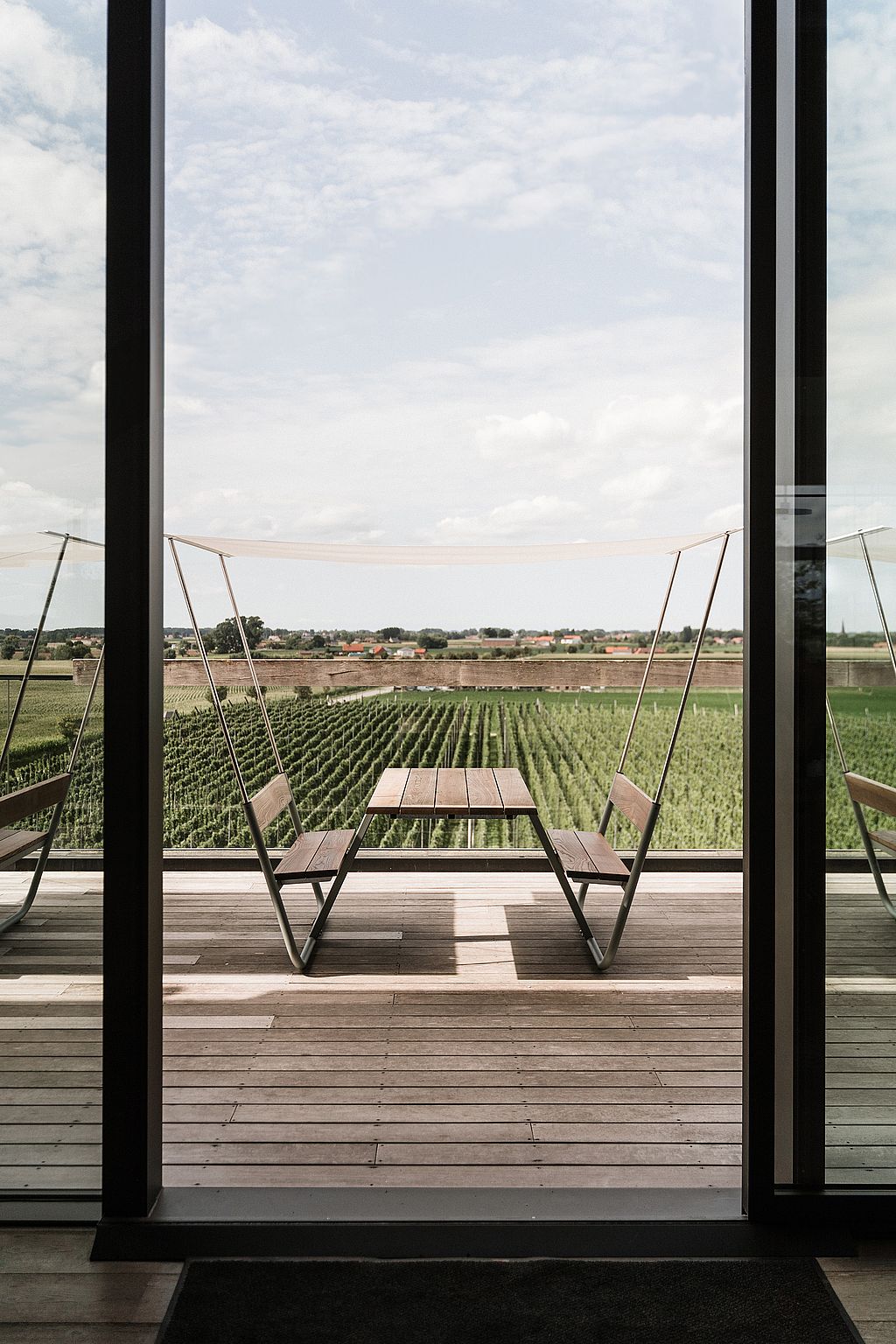 An empty wooden Manille table with integrated benches and a white canopy stands on the terrace, centered against a wide view of the fields.