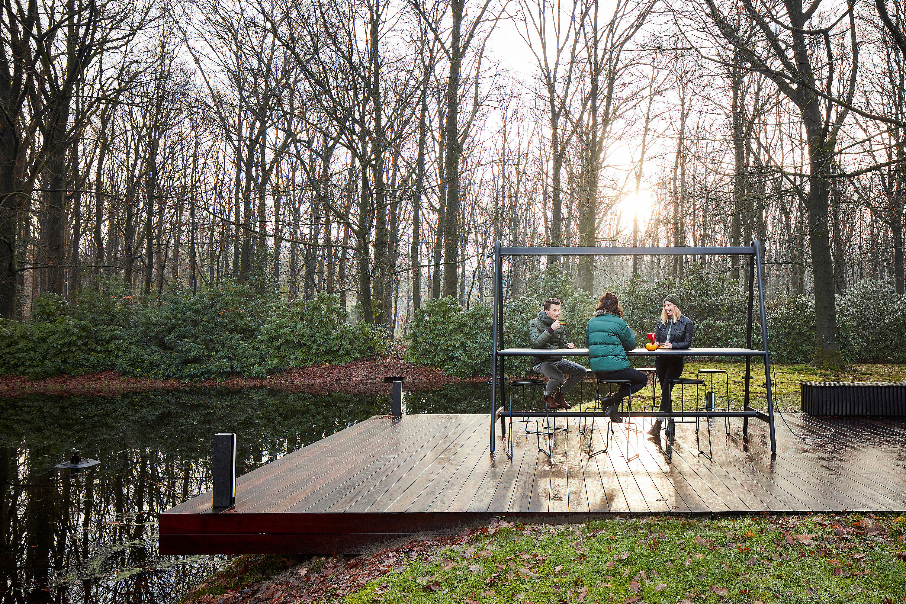 Drei Personen sitzen am AMAi-Tisch auf einer Holzterrasse am Wasser, mit Winterbäumen und tiefstehender Sonne dahinter.