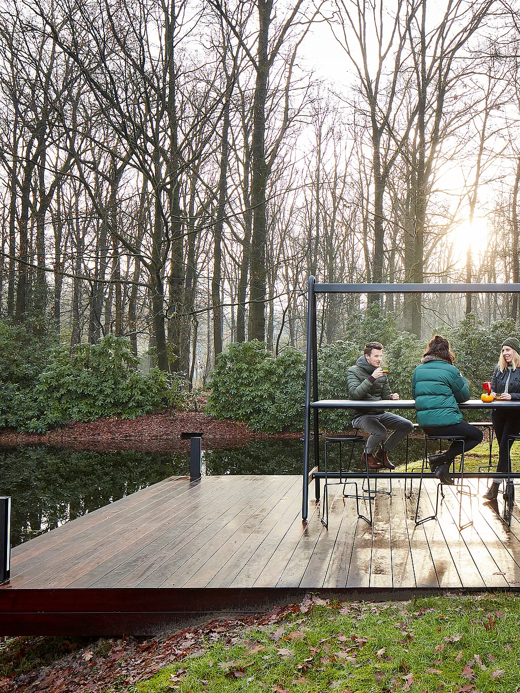 Three people sit at the AMAi table on a wooden deck by the water, with winter trees and low sun behind them.