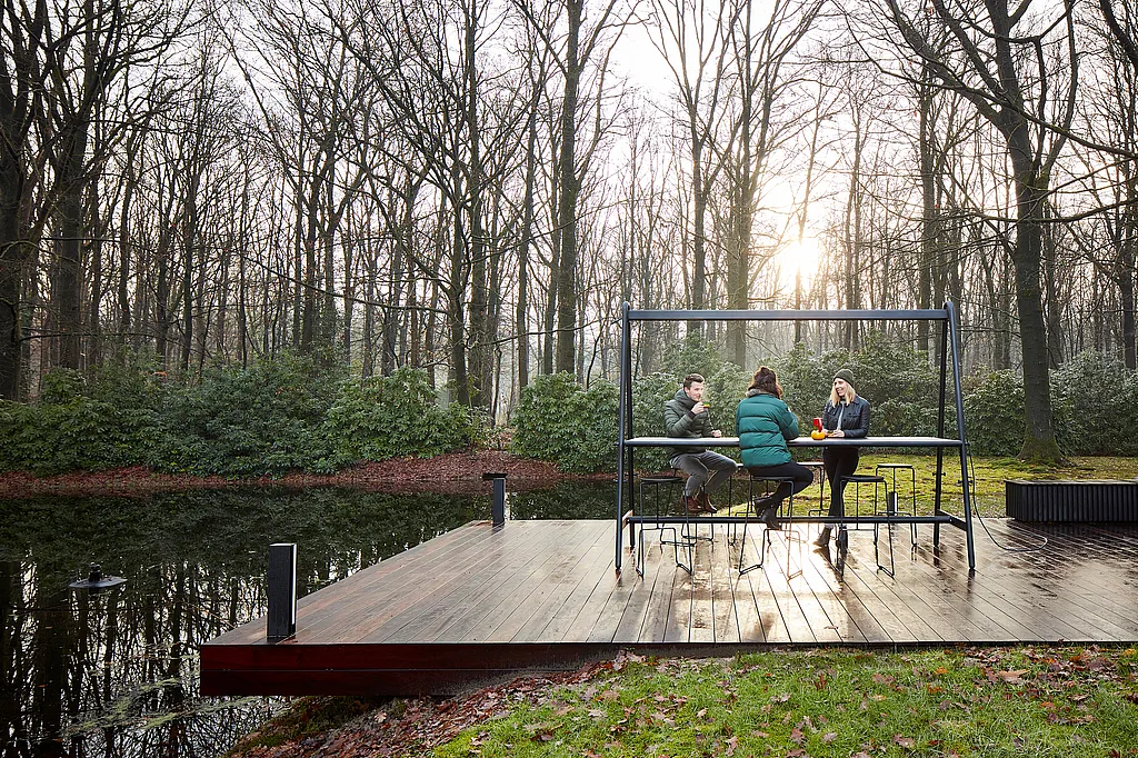 Drei Personen sitzen am AMAi-Tisch auf einer Holzterrasse am Wasser, mit Winterbäumen und tiefstehender Sonne dahinter.