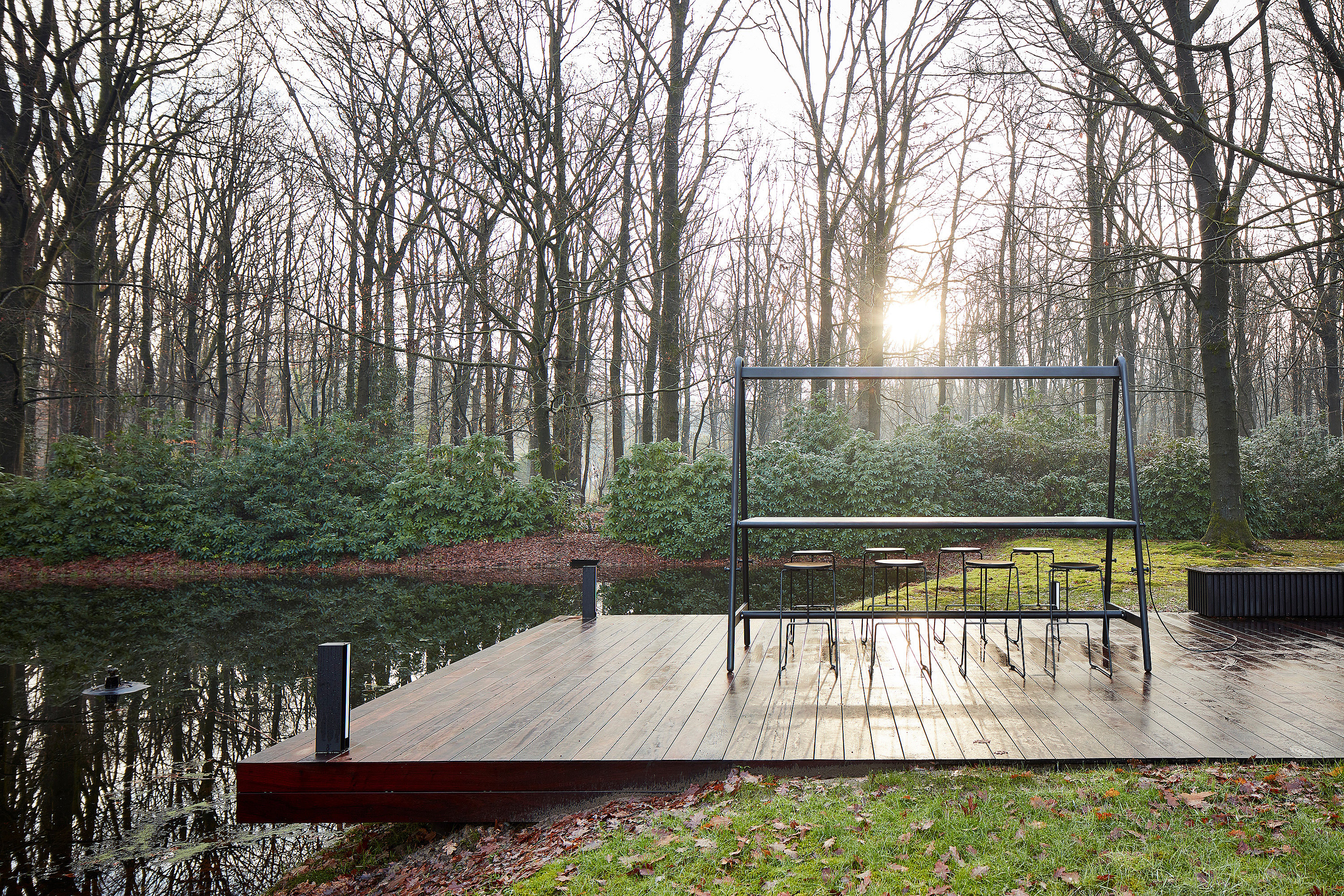 Een lege donkere AMAi hoge tafel met verschillende Tiki krukken staat op een houten dek naast een rustig meer, omlijst door kale winterbomen en laag groen.