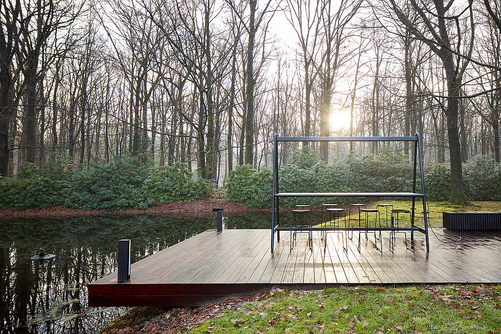 Een lege donkere AMAi hoge tafel met verschillende Tiki krukken staat op een houten dek naast een rustig meer, omlijst door kale winterbomen en laag groen.