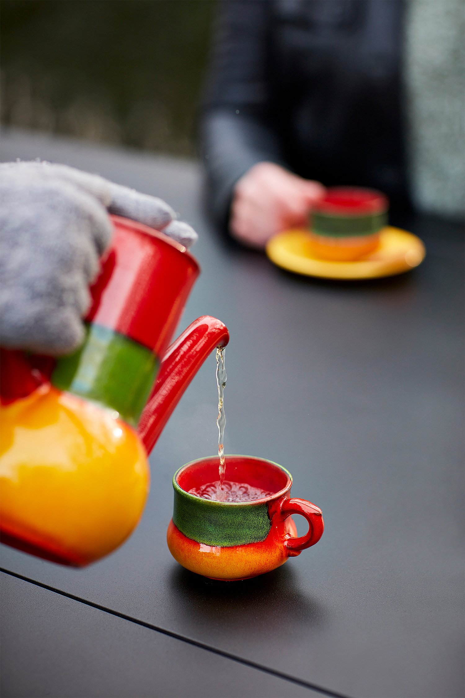 Close-up van thee die van een felrood-gele theepot wordt gegoten in een bijpassende beker op het donkere AMAi-tafelblad.