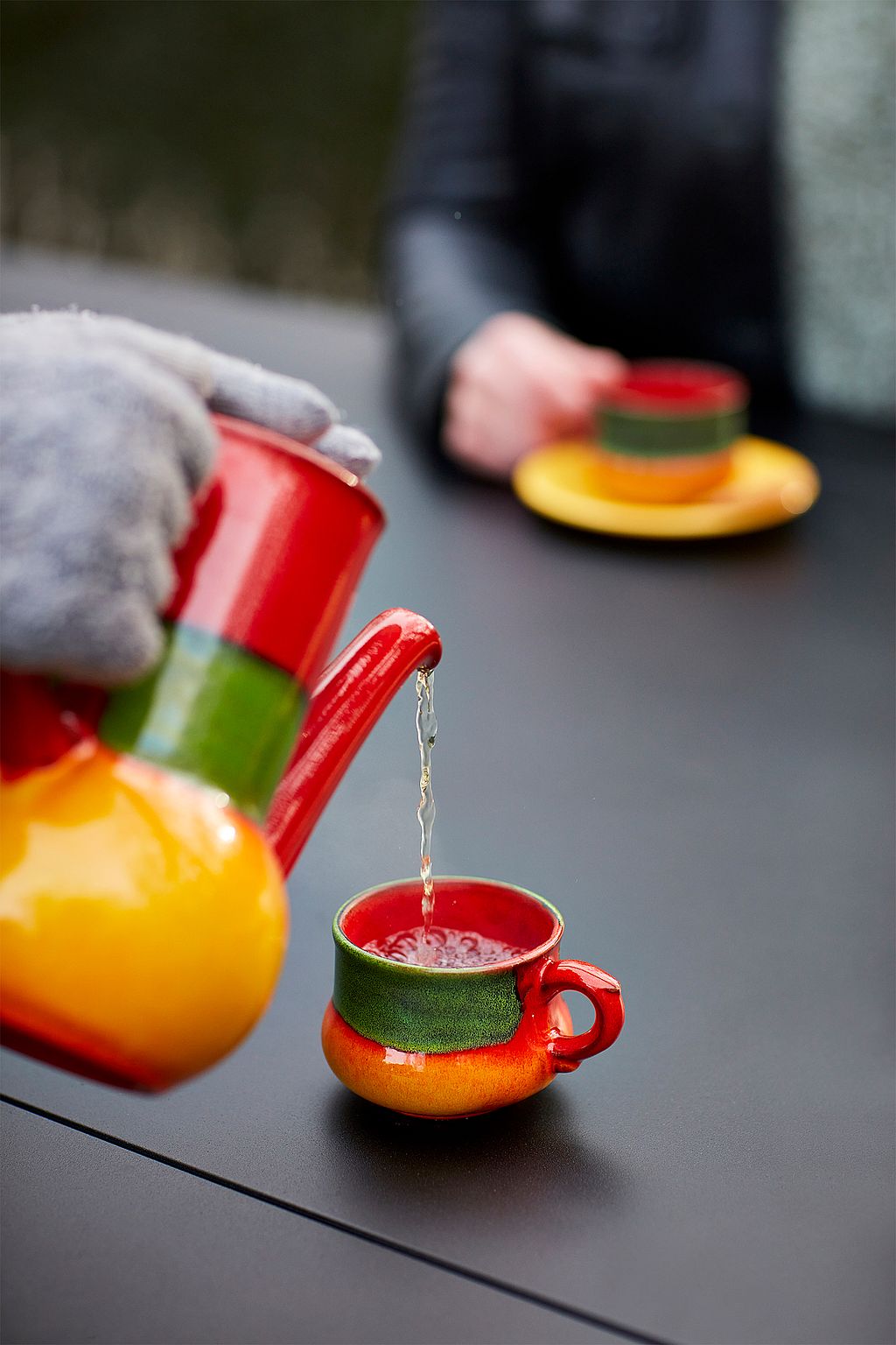 Close-up van thee die van een felrood-gele theepot wordt gegoten in een bijpassende beker op het donkere AMAi-tafelblad.