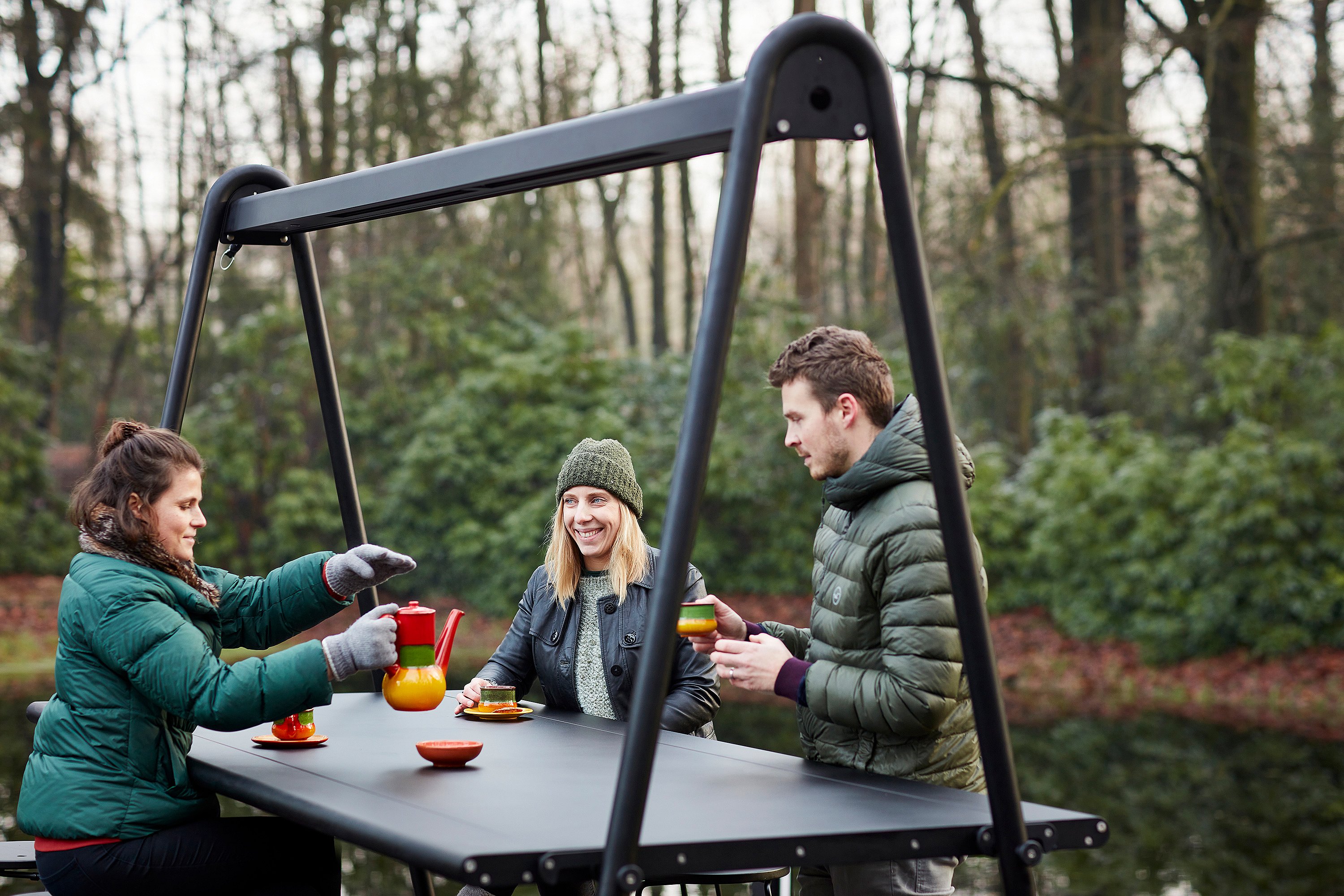 Drie mensen drinken thee aan een donkere AMAi hoge tafel met Tiki krukken op een houten dek naast een meer, omgeven door winterbomen in zacht zonlicht.